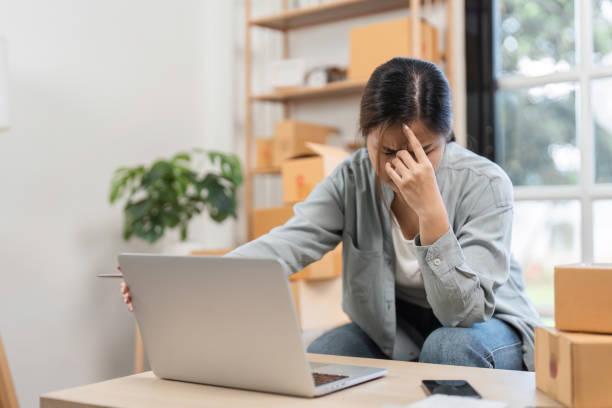 Young Woman Stressed About Online Sales System While Managing E-commerce Business from Home Office A young woman feeling stressed while managing her online sales system from her home office, surrounded by boxes and a laptop. Regulatory challenges stock pictures, royalty-free photos & images